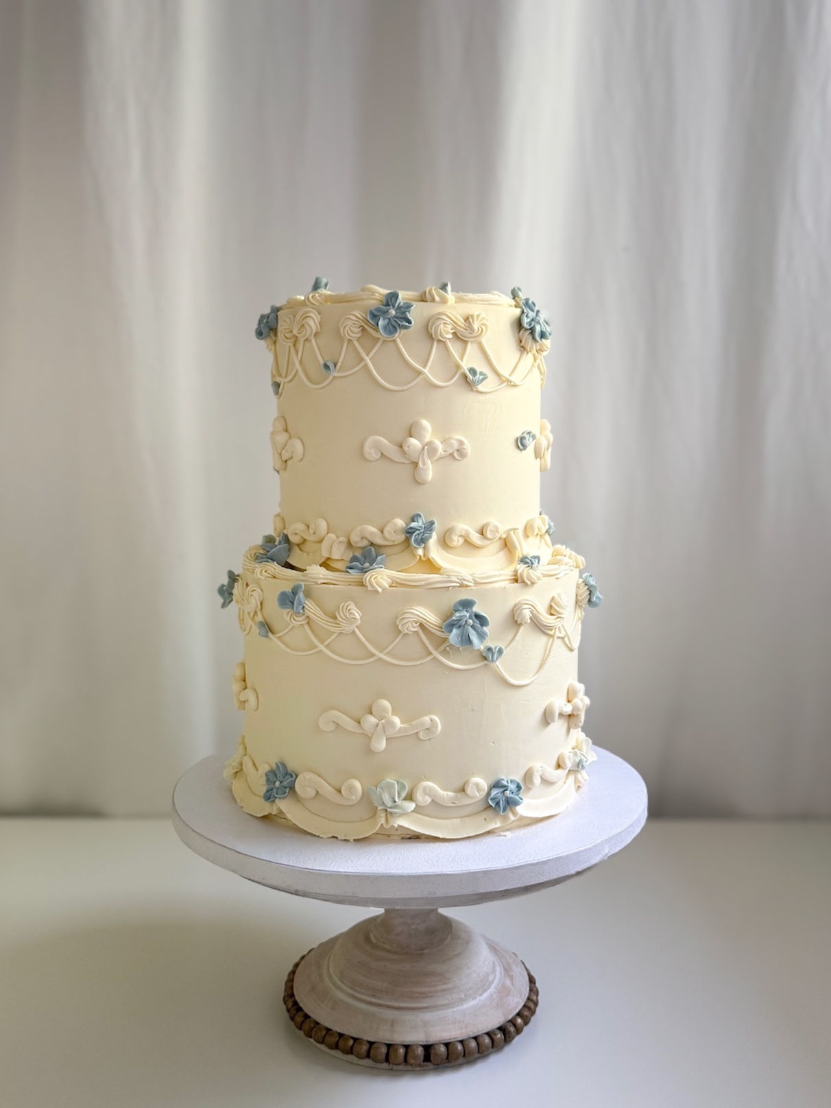 A two-tier cream vintage cake decorated with intricate buttercream piping and small dusty blue flowers on a wooden stand.